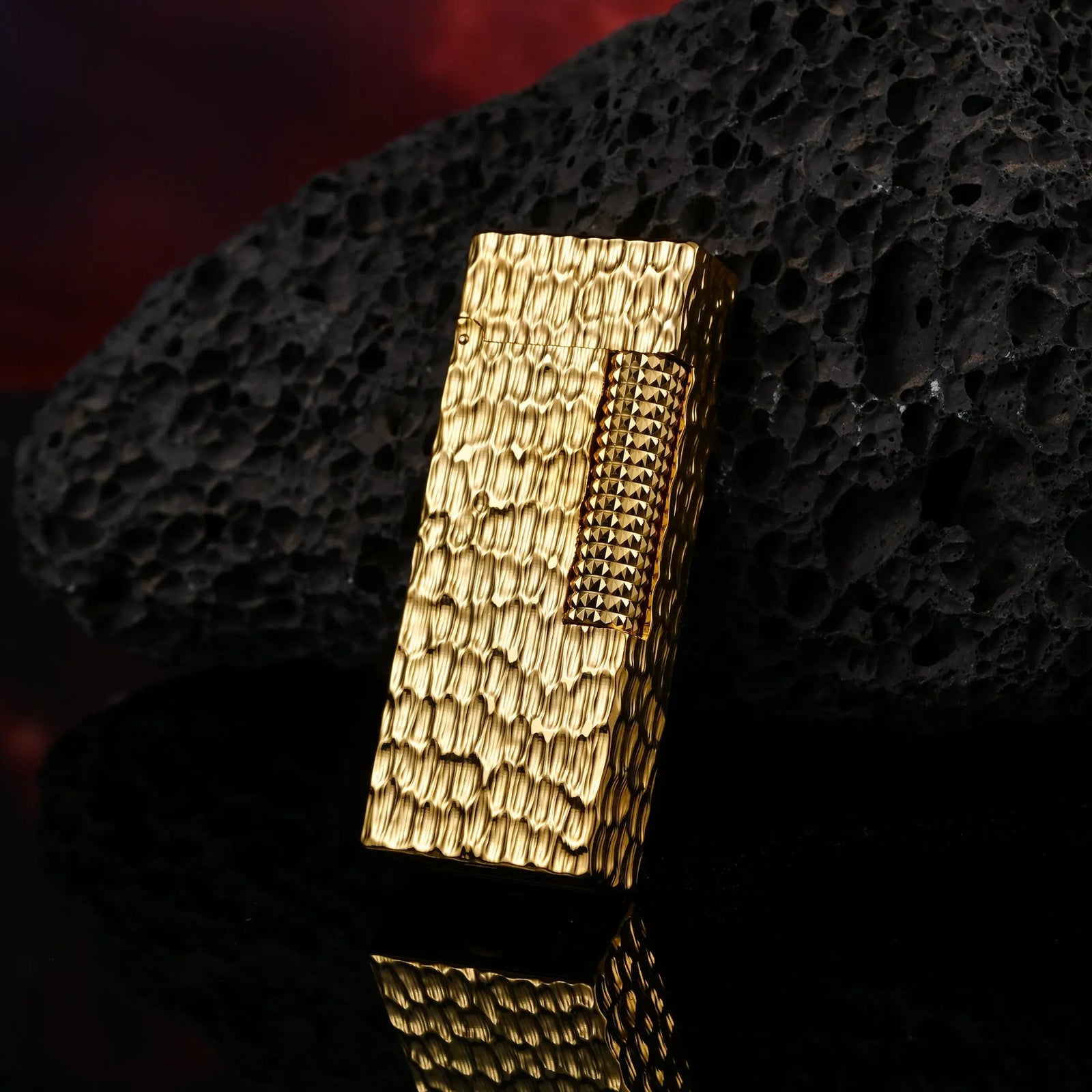 A slim Golden Crater Side Slip lighter featuring a deeply textured hammered gold surface, leaning against a dark volcanic rock with a sharp reflection.