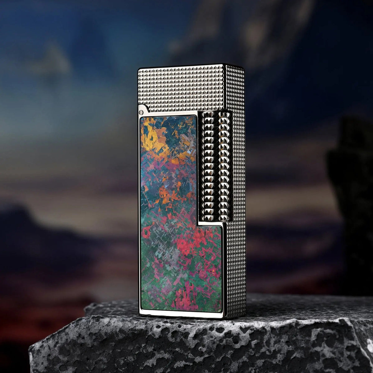 A slim Side Slip lighter featuring a vibrant Iridescent Ti inlay with splashes of pink, gold, and green, standing on a rock against a dramatic twilight mountain background.
