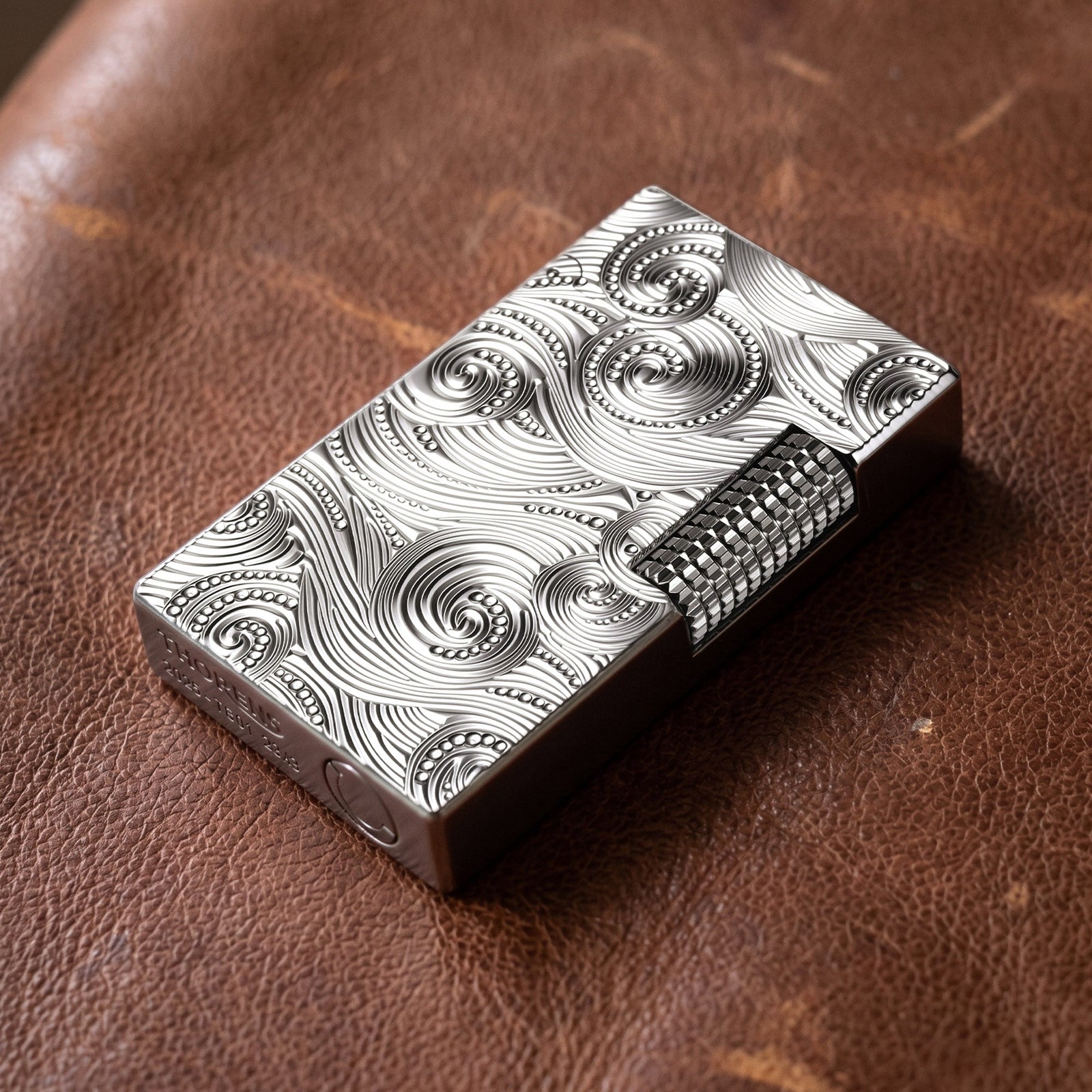 A close-up view of a premium silver Thorens side-slip lighter featuring an intricate, deeply carved vortex pattern on its brass body, showcased on a wooden surface.