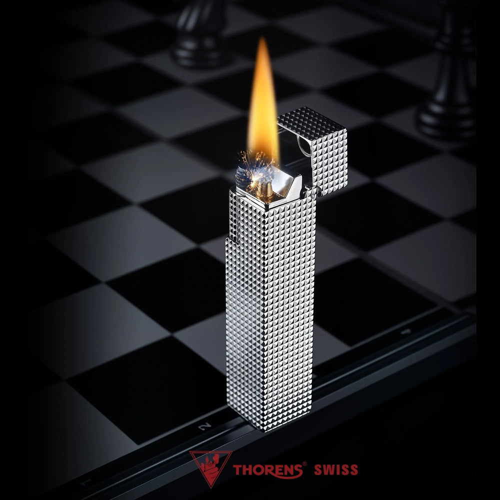 Lit silver small square textured Thorens side-slip rectangular lighter standing open on a black and white checkerboard surface