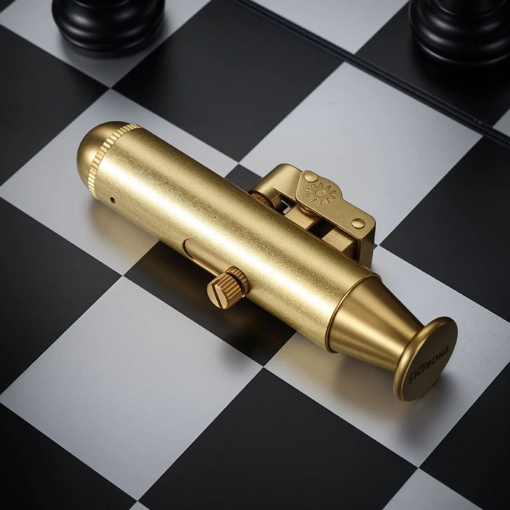 A brushed brass trench lighter in a unique submarine-inspired shape, resting on a black and white chessboard with mechanical adjustment screws and a sun logo engraving.