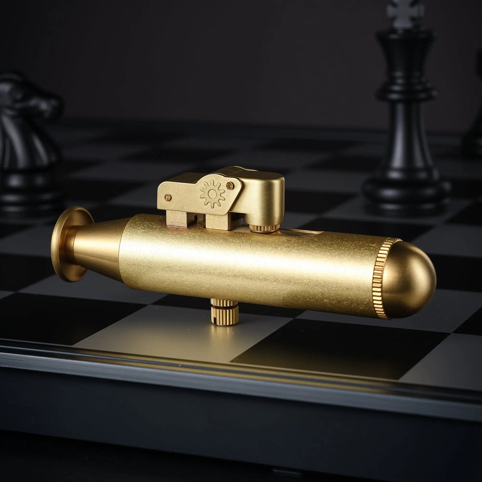 A reverse side view of the Submarine brass trench lighter on a chessboard, showcasing the textured gear detail on the ignition arm and the uniform brushed metal finish.
