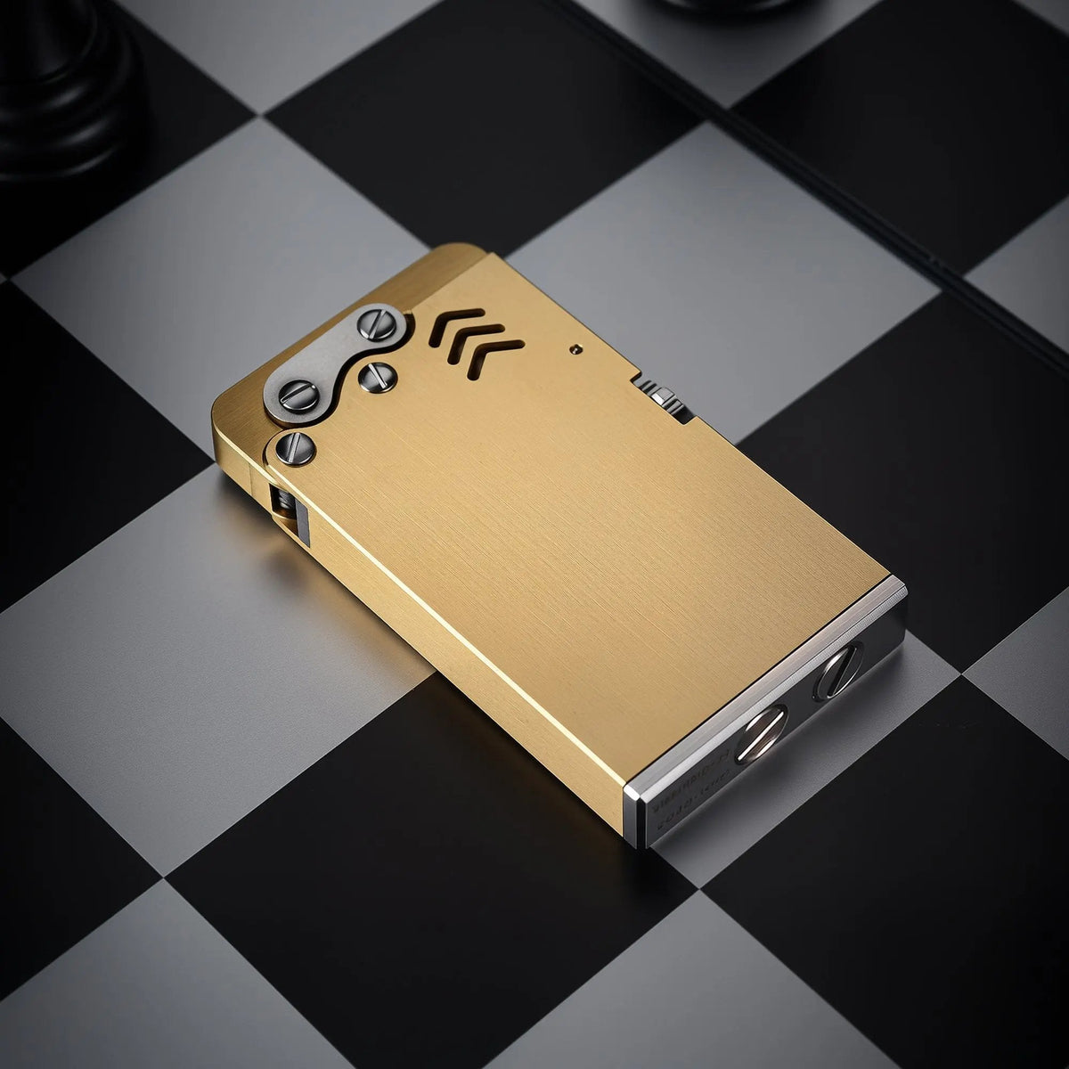 Brushed brass Thorens Thin Ejection lighter with industrial screw details, lying on a black and white checkered chessboard background.