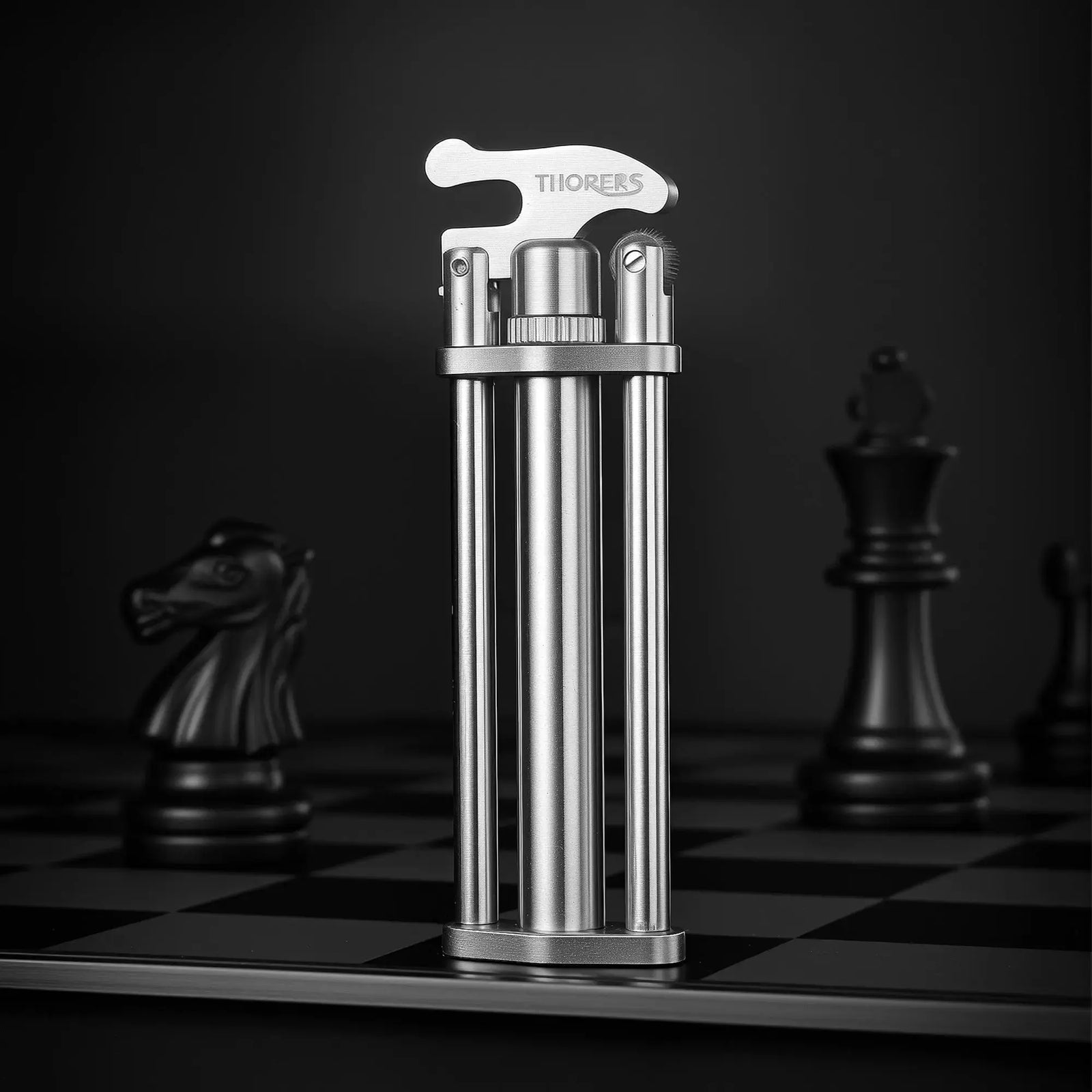 A slim silver three-section stick lighter with a unique triple-column body and engraved Thorens lever, standing prominently on a chessboard beside blurred black chess pieces.