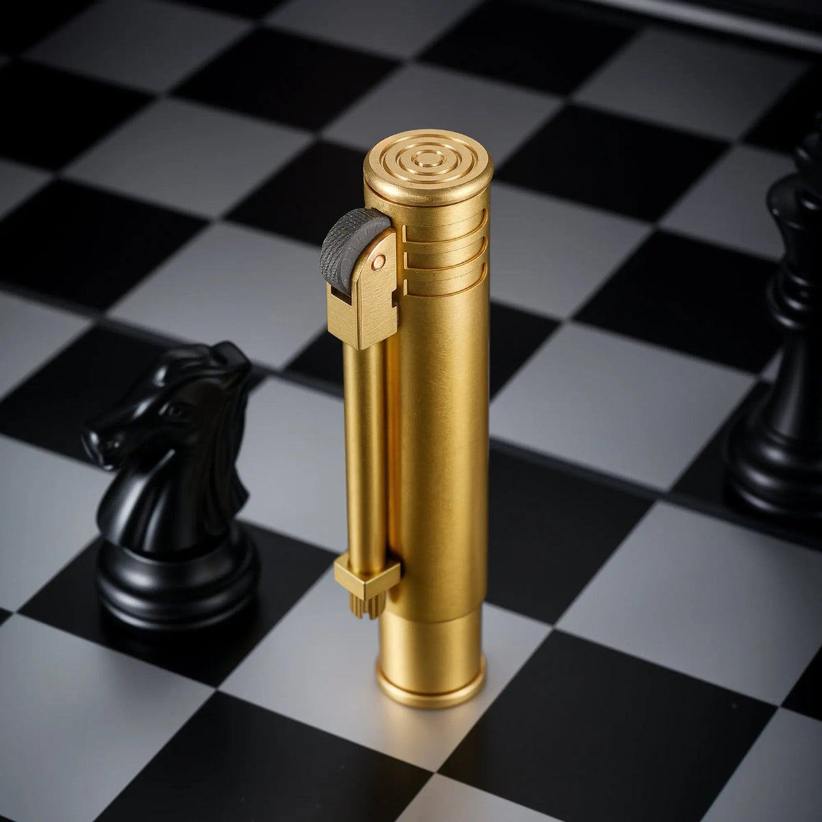 A vertical Torch Stick lighter in a matte brass finish standing on a black and white chessboard next to a black knight chess piece.
