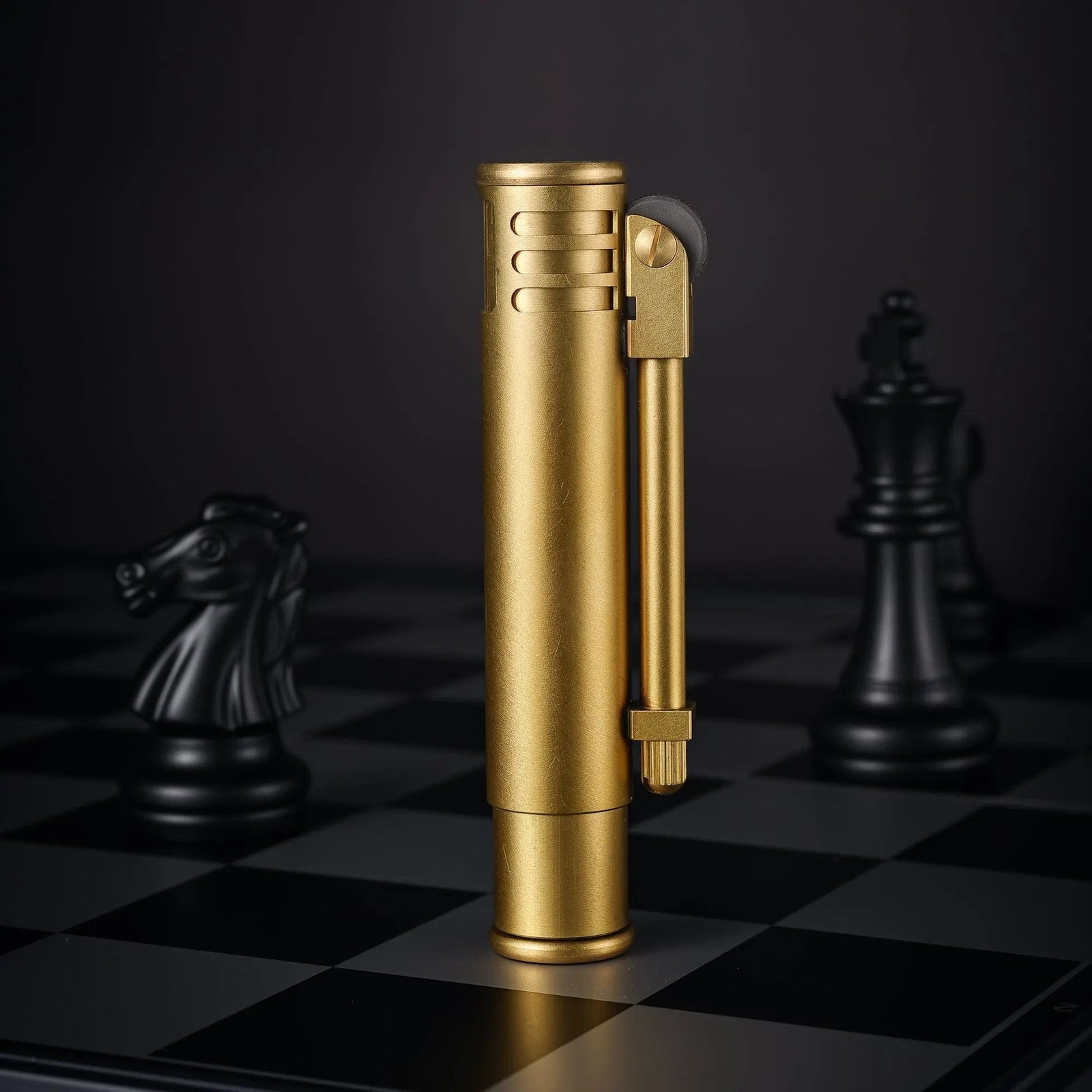 A vertical standing Torch Stick lighter in a textured brass finish, positioned on a chessboard with black chess pieces in the background.