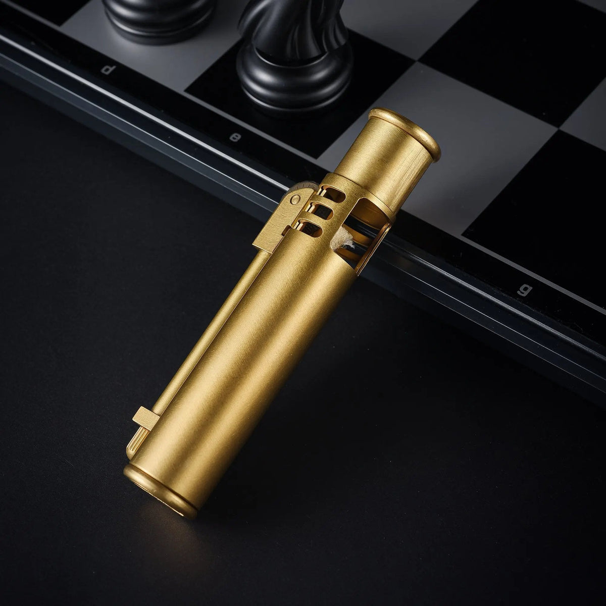 A slim, cylindrical Torch Stick lighter in a brushed brass finish resting on a dark surface next to a black and white chessboard with chess pieces.