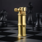 A vertical studio shot of the Mini Bamboo lighter in brushed brass, standing on a silver and black chessboard with out-of-focus black chess pieces in the background.