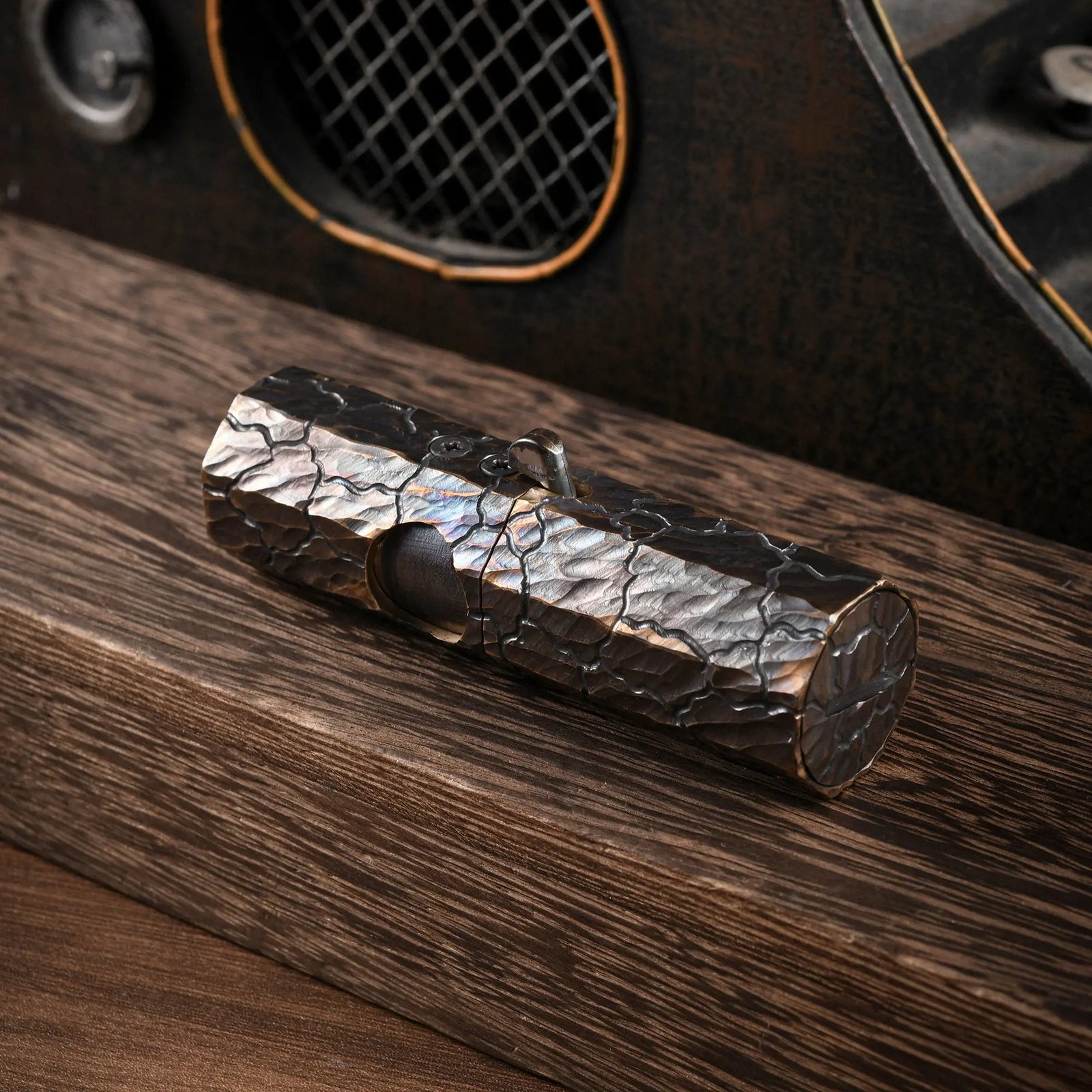 A Thorens Octagon Trigger lighter with a deep hand-carved "Earth Fissure" texture and dark iridescent patina, resting on a rustic wood surface in an industrial setting.